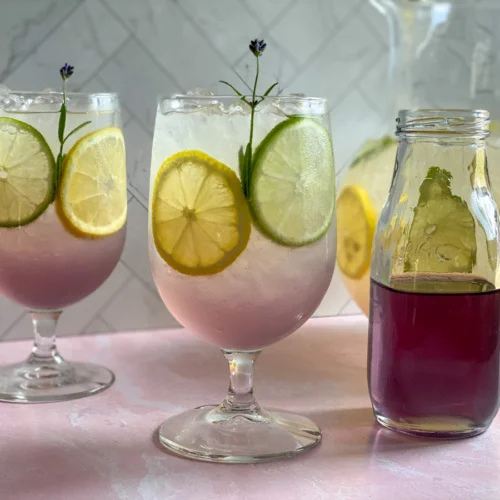 Two glasses with lavender lemonade and a bottle with culinary lavender and blue pea flower syrup