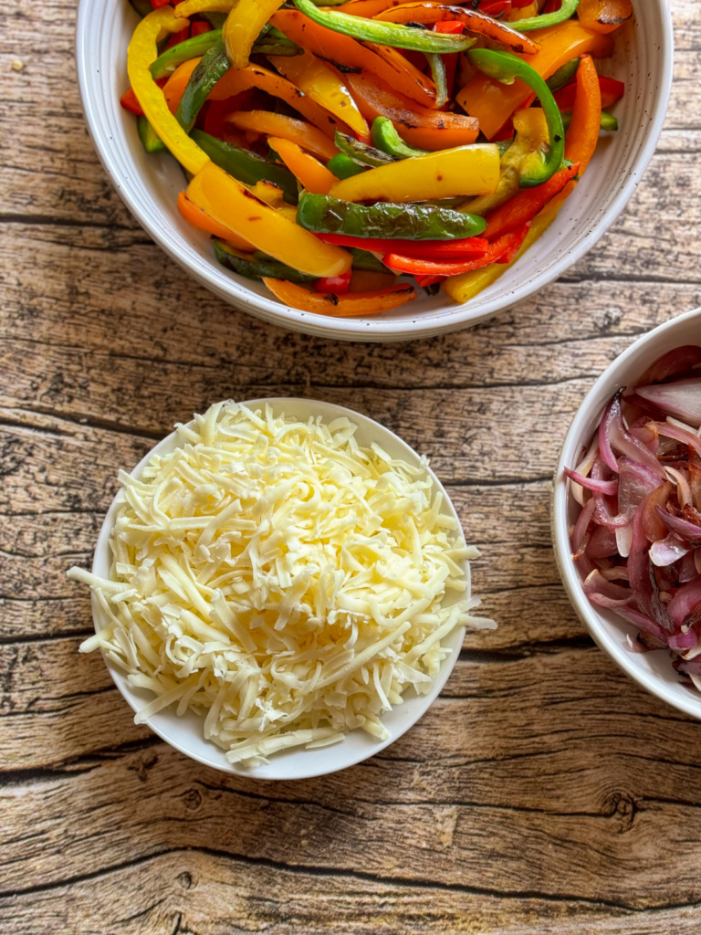 Bowls on a wooden surface contain sliced bell peppers, shredded cheese, and sliced red onions