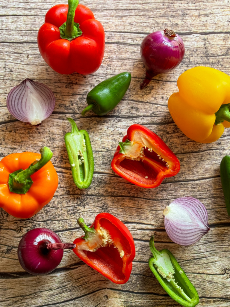 Colorful bell peppers, jalapeños, and red onions, both whole and cut, arranged on a wooden surface