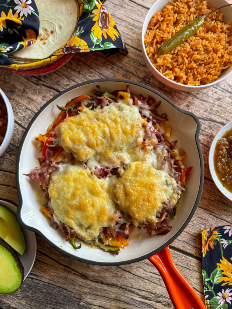 Una sartén con ternera con queso, rodeada de tazones de arroz mexicano, aguacates en rodajas, salsa verde y tortillas envueltas en una servilleta sobre una mesa de madera.