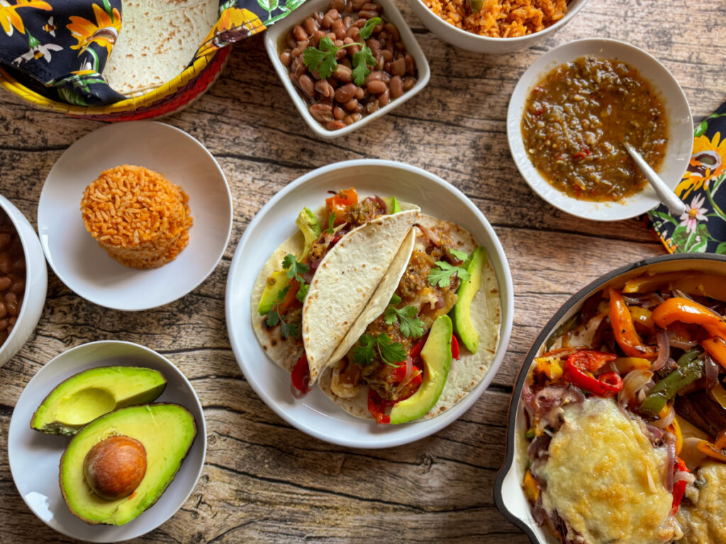 Sobre una mesa de madera se extienden varios platos mexicanos, que incluyen tacos, arroz, frijoles, aguacate y un plato con fajitas de ternera estilo Tex-Mex con queso gratinado.
