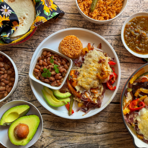 A wooden table set with a variety of Mexican dishes including cheese-topped Tex-Mex fajitas, beans, rice, avocado slices, tortillas in a colorful cloth, and bowls of salsa and sauce.