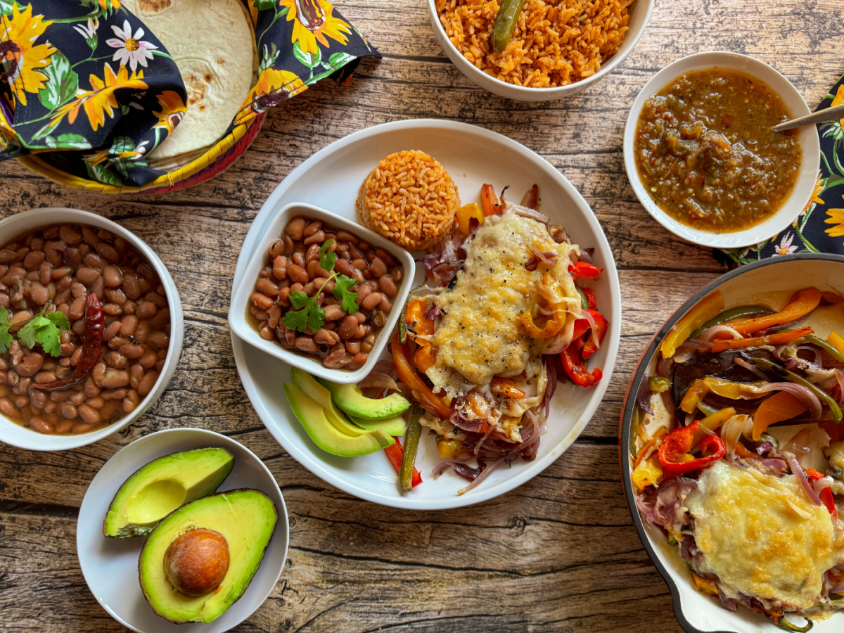 Una mesa de madera con una variedad de platos mexicanos que incluyen fajitas de ternera estilo Tex-Mex cubiertas con queso, frijoles, arroz, rodajas de aguacate, tortillas en un paño colorido y tazones de salsa y salsa.